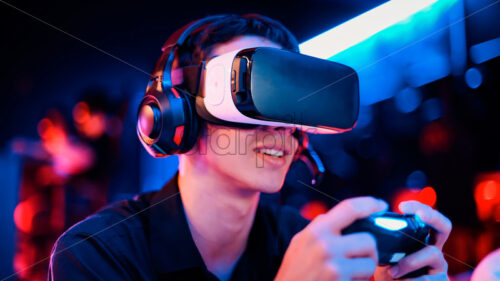 Close view of a smiling teen playing a game console in VR headset and headphones using gamepad. Blue and red illumination - Starpik Stock