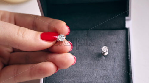 Close view of a woman holding earring with diamonds. A box with another earring behind, hands with red nails - Starpik