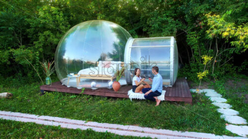 Drone view of a happy couple drinking wine near transparent bubble tent at glamping. Nature around - Starpik Stock