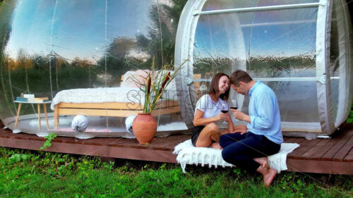 Happy couple drinking wine near transparent bubble tent at glamping. Nature around - Starpik Stock