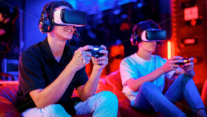 Premium stock photo –Two teen friends are playing a game console in VR headset and headphones using gamepads and smiling while sitting on bean bags. Blue and red illumination - Starpik