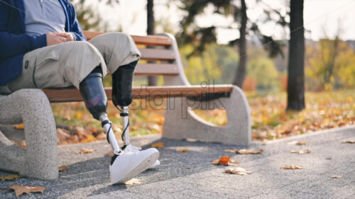 Slow motion view of a man with prosthetic legs and white sneakers. Sitting on a bench in a park. Fallen yellow leaves around - Starpik Stock