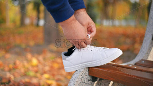 Slow motion view of a man with prosthetic legs and white sneakers. Tying shoelaces with his foot on the bench in a park. Fallen yellow leaves around - Starpik Stock