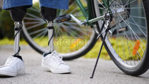 Slow motion view of a man with prosthetic legs. Putting the bike on the kickstand and standing next to it - Starpik Stock