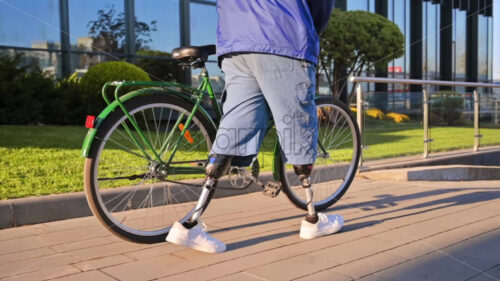 Slow motion view of a man with prosthetic legs. Walking with a bicycle on the street with greenery - Starpik Stock