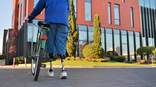 Slow motion view of a man with prosthetic legs. Walking with a bicycle on the street with greenery - Starpik Stock