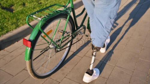 Slow motion view of a man with prosthetic legs. Walking with a bicycle on the street with greenery - Starpik Stock