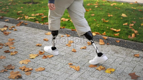 Slow motion view of a walking man with prosthetic legs and white sneakers in a park. Fallen yellow leaves on the ground - Starpik Stock