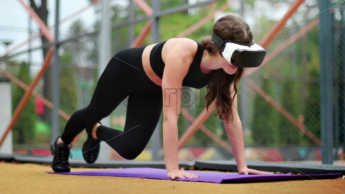 Smiling woman in VR headset in a tracksuit doing exercises on a yoga mat on sports field in a park. Slow motion - Starpik Stock