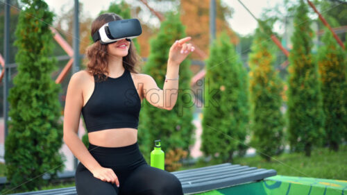Smiling woman using VR headset in a tracksuit while sitting on a bench on a sports field in a park - Starpik Stock