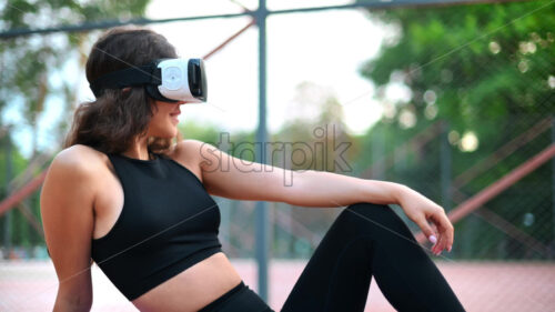 Smiling woman using VR headset in a tracksuit while sitting on yoga mat on a sports field in a park - Starpik Stock