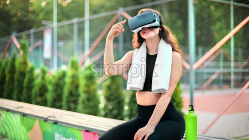 Smiling woman using with towel is using VR headset in a tracksuit while sitting on a bench on a sports field in a park - Starpik Stock