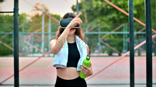 Smiling woman with towel using VR headset in a tracksuit while holding a water bottle on a sports field in a park - Starpik Stock