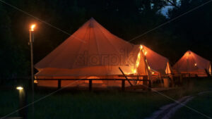 Tents with burning torches, lamps and wooden chairs at glamping, forest around, night - Starpik