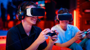 Two teen friends are playing a game console in VR headset and headphones using gamepads and smiling while sitting on bean bags. Blue and red illumination - Starpik Stock