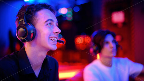 Two teen friends in headphones are playing a game console using gamepads and smiling while sitting on bean bags. Blue and red illumination - Starpik Stock