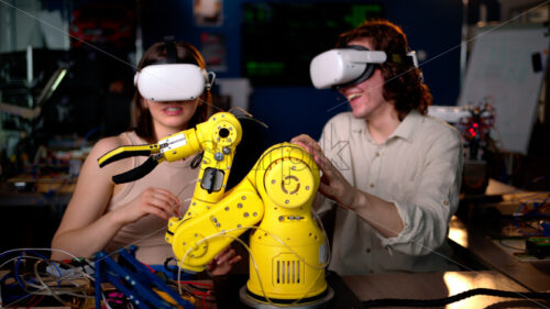 Two young engineers fixing a yellow robot arm in the workshop, wearing VR virtual reality headset - Starpik Stock