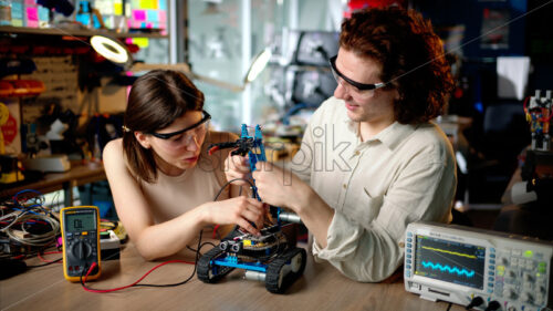 Two young happy engineers fixing a mechanical robot car in the workshop, using VR virtual reality headsets - Starpik Stock