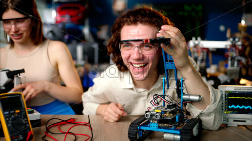 Two young happy engineers fixing a mechanical robot car in the workshop, using VR virtual reality headsets - Starpik Stock