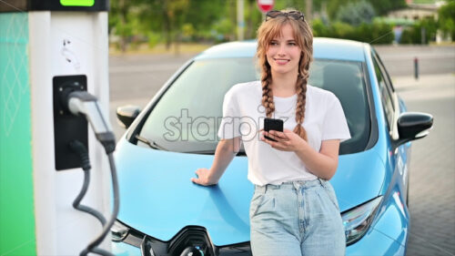 Video – A young blonde woman using smartphone at a car charging station with charging electric car nearby. Chisinau, Moldova. Slow motion - Starpik Stock