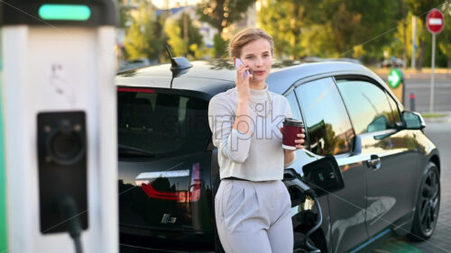 Video – A young blonde woman with coffee talking on the smartphone at a car charging station with electric car nearby in Chisinau at sunset, Moldova - Starpik Stock