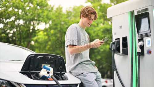 Video – A young man using smartphone at a car charging station with charging electric car nearby. Chisinau, Moldova. Slow motion - Starpik Stock