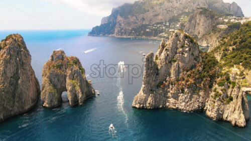 Video – Aerial drone cinematic view of the Tyrrhenian sea coast of Capri, Italy. Rocky cliffs, blue water, floating boats, greenery, town in the distance - Starpik Stock