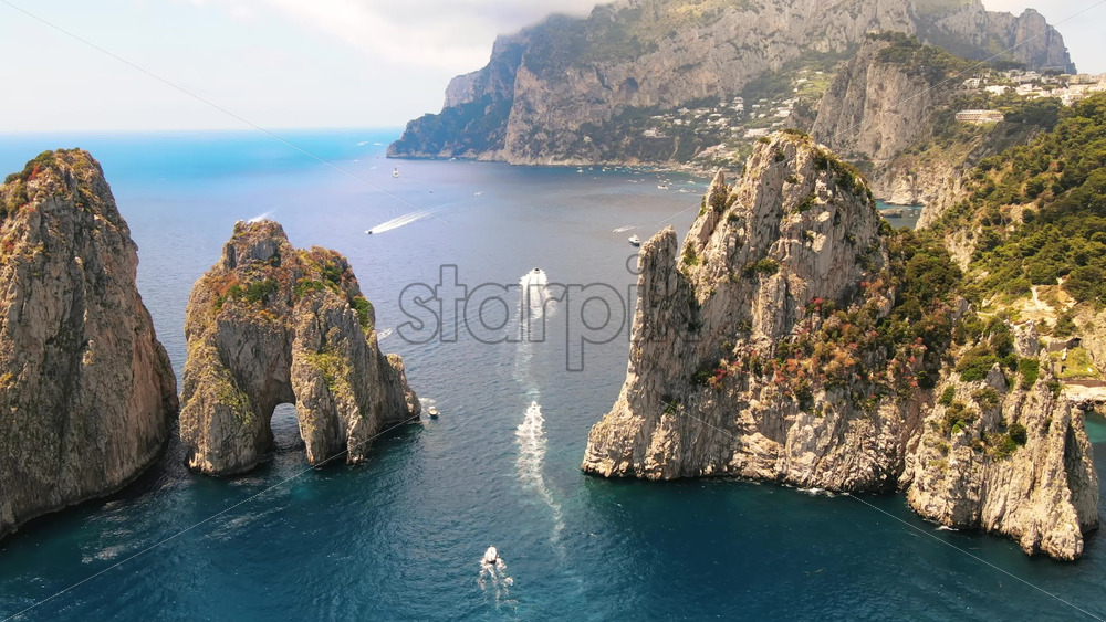 Video - Aerial drone cinematic view of the Tyrrhenian sea coast of Capri, Italy. Rocky cliffs, blue water, floating boats, greenery, town in the distance