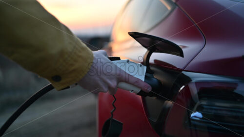 Video – Close view of a man plugging charging cable into a parked red electric car at sunset - Starpik Stock