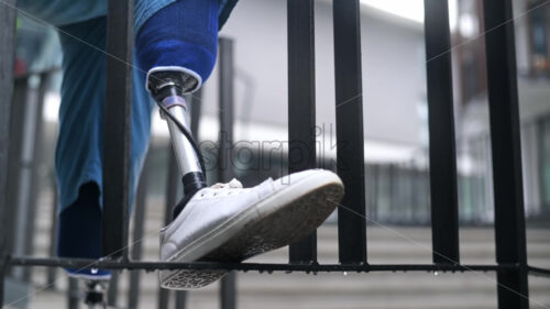 View of a man with prosthetic legs and white sneakers at rainy weather. Rested one foot on the fence - Starpik Stock