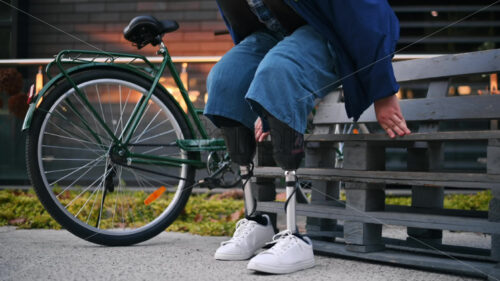 View of a man with prosthetic legs. Getting up from the bench and leaving with his bicycle - Starpik Stock