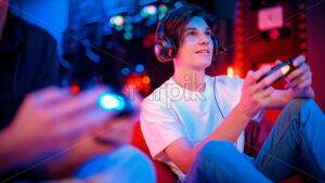 View of a smiling teen playing a game console in headphones using gamepad while sitting on bean bag. Blue and red illumination - Starpik Stock