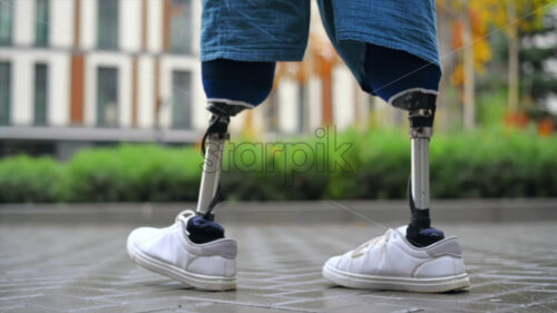 View of a walking man with prosthetic legs and white sneakers at rainy weather, greenery on the background - Starpik Stock