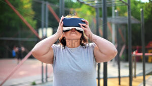 Woman with overweight in gray t-shirt and leggings exercising on a sports field in a park while wearing a virtual reality headset. Fitness lifestyle - Starpik Stock