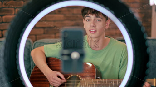 Young man playing on a guitar and singing, shooting himself on smartphone on a tripod with ring light in a studio - Starpik Stock