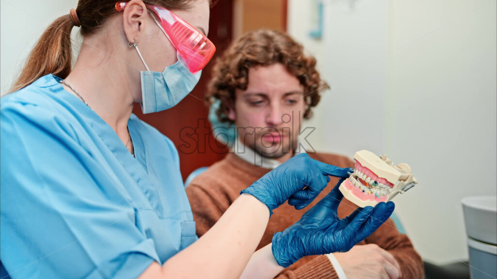 Video Dentist wearing a protective mask, goggles, and gloves showing a teeth mock up to a patient in a dental cabinet