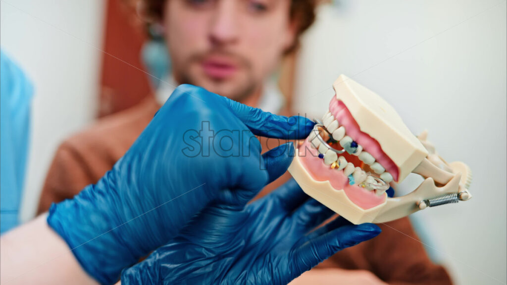Video Dentist wearing a protective mask, goggles, and gloves showing a teeth mock up to a patient in a dental cabinet