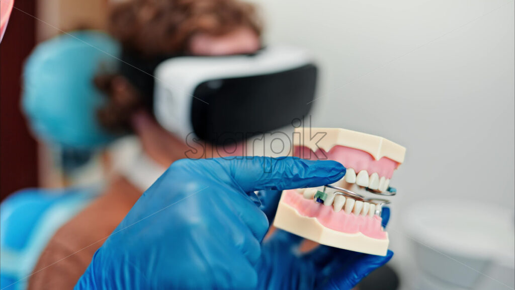 Video Dentist wearing a protective mask, goggles, and gloves showing a teeth mock up while the patient is wearing a Virtual Reality headset in dental cabinet