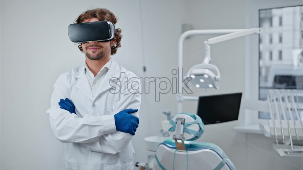 Video Dentist wearing a scrub and a Virtual Reality headset while standing and smiling in a dental cabinet, in front of a window