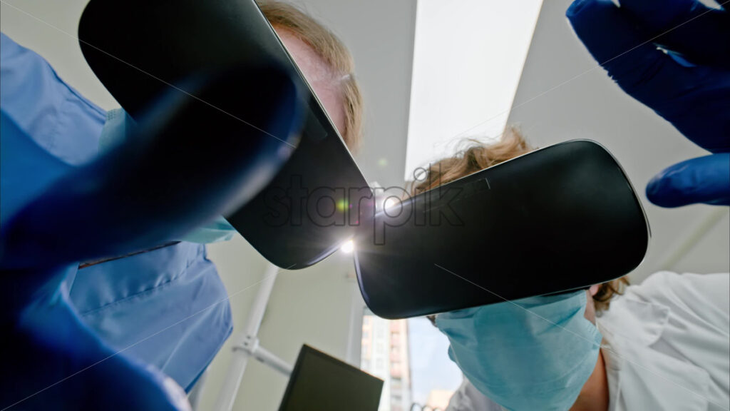 Video Low angle view of two dentists wearing protective masks, gloves and Virtual Reality headsets gesturing under bright procedural lights