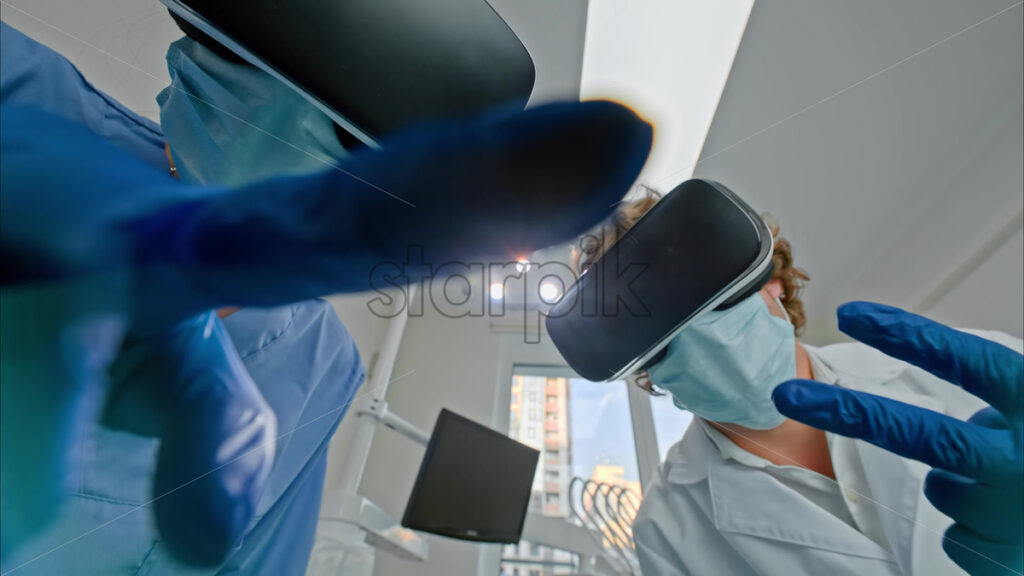 Video Low angle view of two dentists wearing protective masks, gloves and Virtual Reality headsets gesturing under bright procedural lights