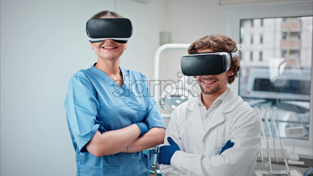 Video  Two dentists wearing scrubs and Virtual Reality headsets while standing and smiling in a dental cabinet, in front of a window