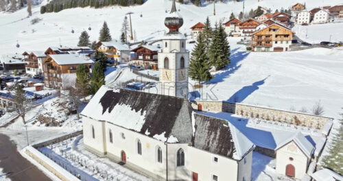 Aerial drone view of the Parrocchia di Colfosco in the Colfosco mountain village covered in snow, in South Tyrol, Dolomites, Northern Italy - Starpik Stock