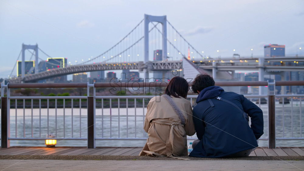 A man and a woman couple sitting in front of a view of the Rainbow Bridge and the skyline of Tokyo, Japan in the evening - Starpik Stock
