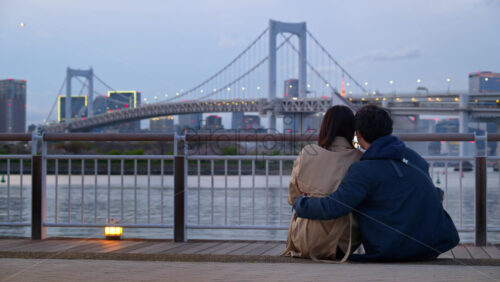 A man and a woman couple sitting in front of a view of the Rainbow Bridge and the skyline of Tokyo, Japan in the evening - Starpik Stock