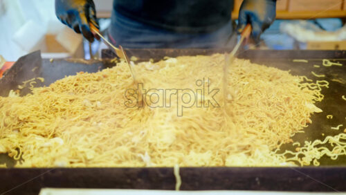 Close up of a vendor using metal spatulas to toss the fried noodles around on the teppan at a street food market in Japan - Starpik Stock