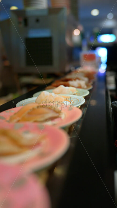 Close up of plates with sushi placed on a rotating conveyor belt moving through the restaurant. Vertical - Starpik Stock