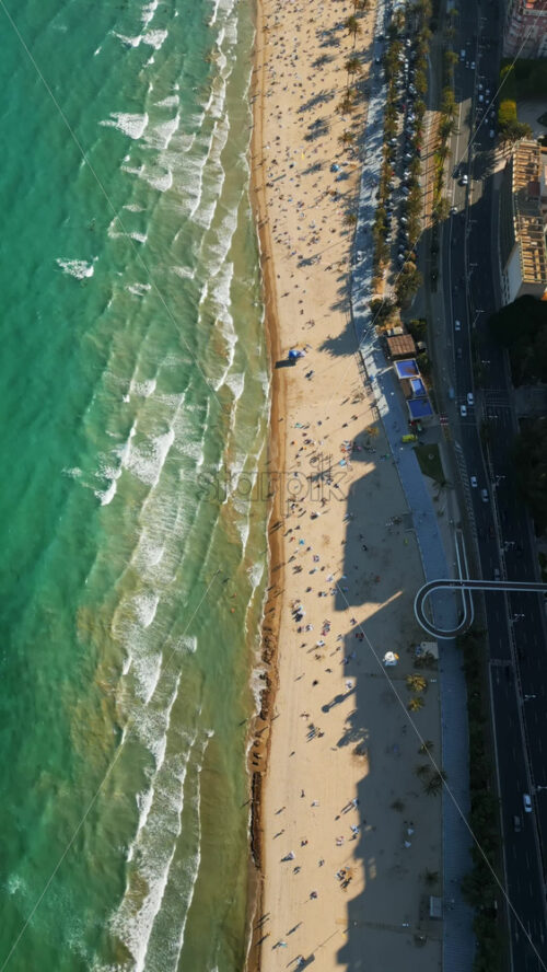 Aerial drone view of cars moving along the coastline with people relaxing on the beach in Alicante, Spain in daylight. Vertical - Starpik Stock