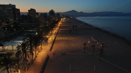 Aerial drone view of people walking on a promenade lined with palm trees near the beach in Alicante, Spain in the evening - Starpik Stock