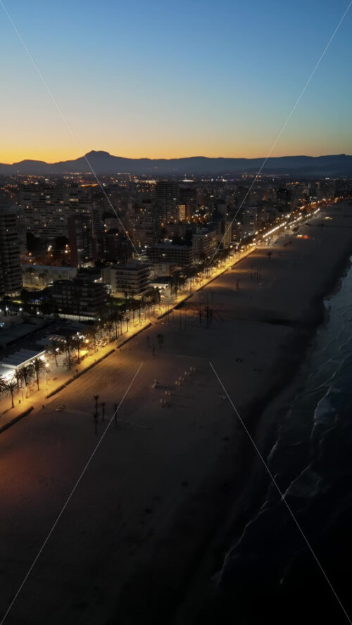 Aerial drone view of people walking on a promenade lined with palm trees near the beach in Alicante, Spain in the evening. Vertical - Starpik Stock